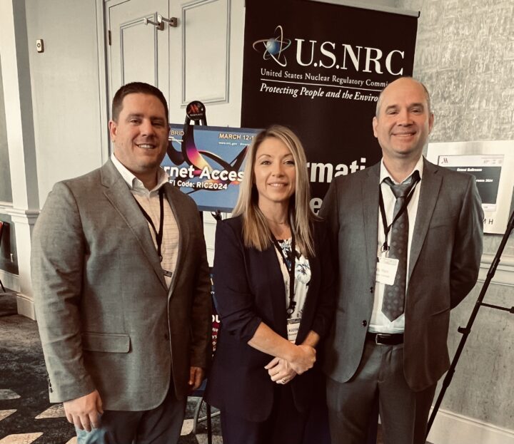 Three professionals from Accelerant Solutions standing in front of a U.S. NRC sign at the 36th Regulatory Information Conference, showcasing their engagement in nuclear industry discussions.