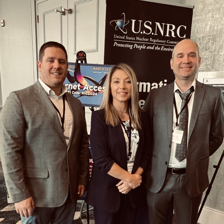 Three professionals from Accelerant Solutions standing in front of a U.S. NRC sign at the 36th Regulatory Information Conference, showcasing their engagement in nuclear industry discussions.